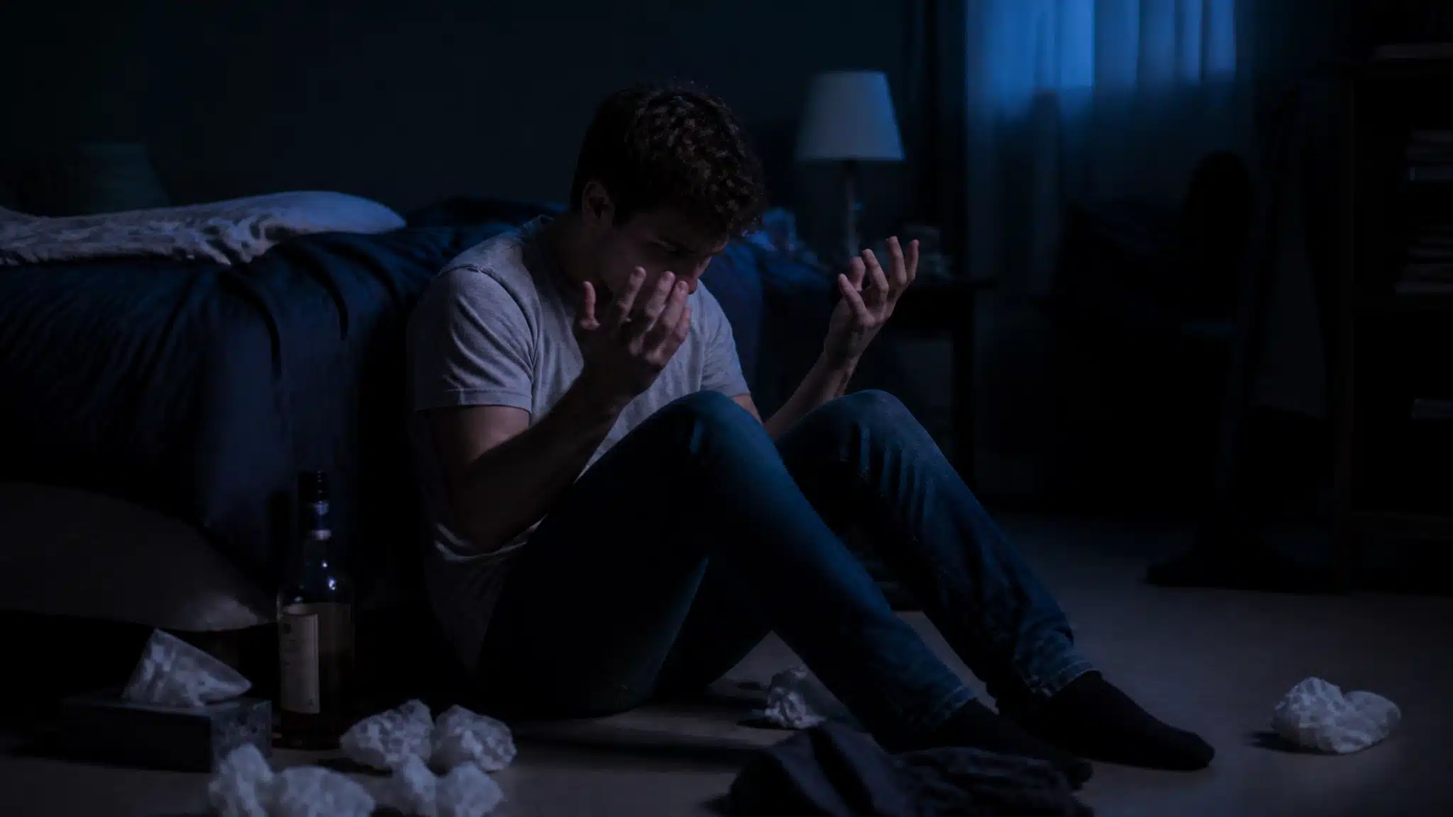 Young man sitting distressed on the floor beside a liquor bottle